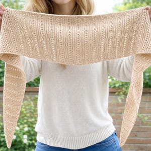 Può includere: Una sciarpa triangolare in maglia di colore beige chiaro caratterizzata da un motivo testurizzato a colonne verticali a coste intervallate da delicati dettagli in pizzo traforato. La sciarpa ha estremità affusolate e una trama uniforme.