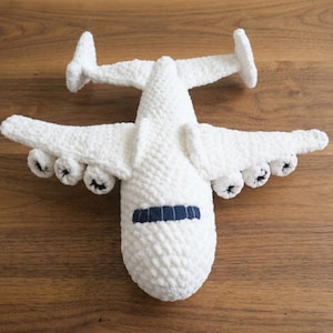 Pode incluir: Um brinquedo de avião em crochê branco com uma superfície de fio texturizado, uma janela de cockpit azul-escura e seis detalhes de motor sob as asas. A aeronave possui uma grande envergadura e uma seção de cauda em forma de T, com toque macio.