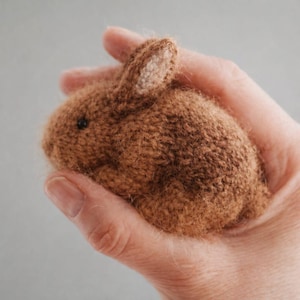 May include: A small, handmade brown knitted bunny with a light-colored inner ear. The bunny is held in a person's hand, showcasing its soft texture and detailed craftsmanship. The bunny's eyes are small and black.
