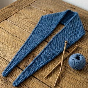 Può includere: Una sciarpa blu lavorata a maglia di forma triangolare, appoggiata su una superficie di legno. Due ferri da maglia in legno e un gomitolo di lana blu sono accanto alla sciarpa. La sciarpa è realizzata in tessuto a maglia.