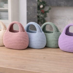 Könnte beinhalten: Fünf kleine, abgerundete Häkelkörbe mit integrierten Griffen, die in einer Reihe angeordnet sind. Die Körbe sind in den Farben Creme, Altrosa, Hellblau, Salbeigrün und Lavendel gehalten. Jeder Korb weist ein dichtes, strukturiertes Maschenbild auf.