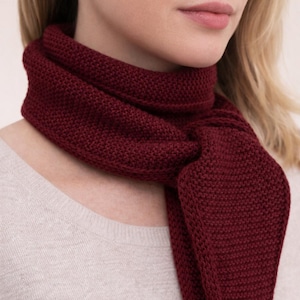 May include: A deep burgundy scarf featuring a dense, textured knit pattern. The scarf is wrapped around the neck and tied in a simple knot, showcasing the consistent, uniform stitch detail throughout the fabric of the accessory.