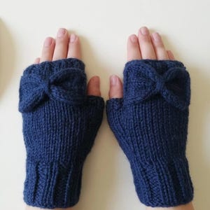 Op de afbeelding: Een paar donkerblauwe vingerloze handschoenen met een gebreide textuur en een opvallende strik op de rug van elke hand. De handschoenen hebben een geribbelde boord bij de pols en een opening voor de duim voor een nauwsluitende pasvorm.