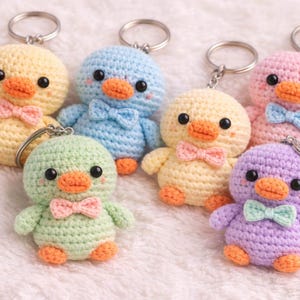May include: Six crocheted duck keychains in various pastel colors including yellow, blue, light green, pink, and purple. Each duck features black bead eyes, an orange beak, orange feet, and a small bow tie in a contrasting color attached to the neck.