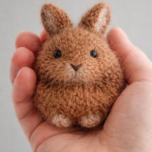 May include: A small, round, brown knitted rabbit figurine with a fuzzy texture, two upright ears, two black bead eyes, and a small stitched nose. The rabbit has light tan paws visible at the base and a soft, dense yarn construction throughout.