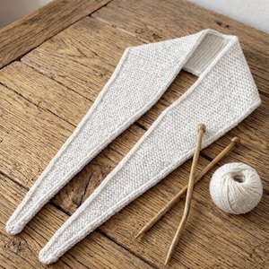 Può includere: Una sciarpa lavorata a maglia grigio chiaro di forma triangolare, appoggiata su una superficie di legno. Due ferri da maglia in legno e un gomitolo di lana sono accanto alla sciarpa. La sciarpa ha un aspetto strutturato.