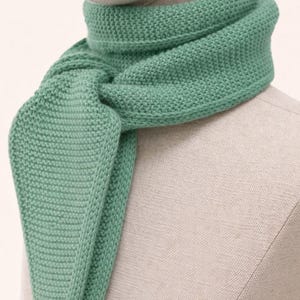 May include: A sage green knitted scarf with a textured garter stitch pattern. The scarf is wrapped around the neck of a mannequin, featuring a triangular pointed end that hangs down in the front, showcasing a consistent knit construction.