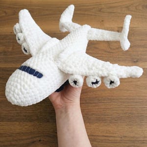 Puede incluir: Un avión de juguete tejido a ganchillo en color blanco con un acabado de hilo suave y texturizado. La aeronave tiene una ventana de cabina azul oscuro y tres motores distintos bajo cada ala, con una cola en forma de T en el fuselaje.