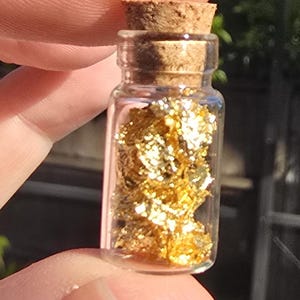 May include: A small, clear glass bottle filled with gold flakes, sealed with a cork stopper. The bottle is held between fingers, with sunlight illuminating the gold contents. The background is blurred, suggesting an outdoor setting.