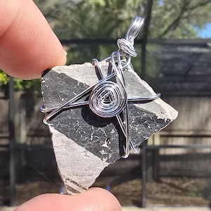 May include: A handmade pendant featuring a piece of black and gray stone wrapped in silver wire. The stone has a geometric pattern and is held in place by the wire, which forms a decorative bail.