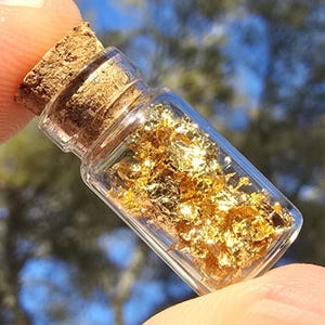May include: A small, clear glass bottle filled with gold flakes, sealed with a cork stopper. The bottle is held between fingers, with a blurred background of blue and green.