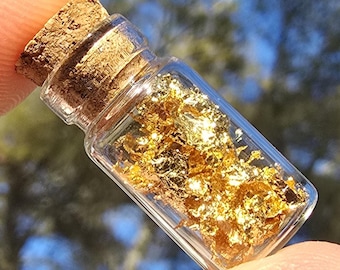 May include: A small, clear glass bottle filled with gold flakes, sealed with a cork stopper. The bottle is held between fingers, with a blurred background of blue and green.