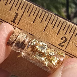 May include: A small, clear glass vial filled with gold flakes, sealed with a cork stopper. The vial is held near a wooden ruler, showing measurements in inches. The gold flakes shimmer, catching the light.