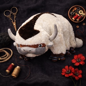 May include: A plush toy of a white and brown bison-like creature with gray horns and facial features. The toy has blue and brown accents on its feet. The image includes sewing supplies and red flowers, suggesting a craft or handmade theme.