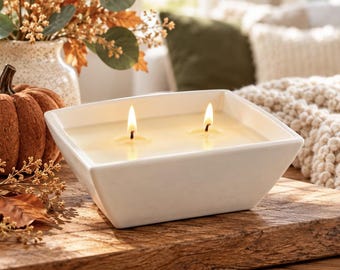 May include: A square white ceramic bowl candle featuring two lit wicks with a smooth, off-white wax surface. The container has a clean, minimalist design with tapered sides, providing a steady glow from the two flames centered in the wax.