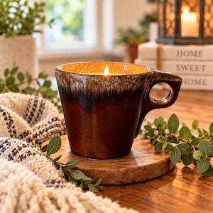 May include: A ceramic mug candle featuring a reactive glaze finish in shades of deep brown and cream. The mug has a wide, rounded handle and contains a lit candle with a single flame. The exterior displays a smooth, glossy texture with a dark drip effect.