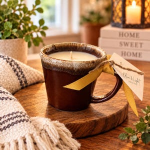 May include: A brown ceramic mug candle with a white wax fill and a single wick. The rim features a textured, lighter brown drip glaze. A mustard yellow ribbon is tied around the handle, holding a white tag printed with Olive & Light Candle Co.