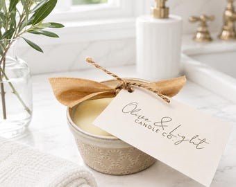 May include: A beige ceramic bowl candle filled with cream-colored wax, tied with a tan ribbon and a rectangular paper tag featuring the black text Olive & Light CANDLE CO. The candle sits on a white marble surface next to a white folded towel.