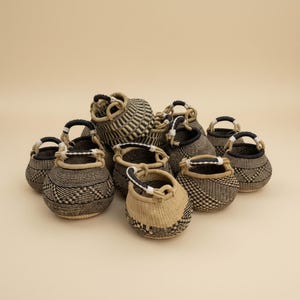 May include: A collection of small, round woven baskets featuring black and natural tan fibre patterns. Each basket has a single arched handle with black and white accents. The baskets are arranged in a cluster, displaying various geometric designs.