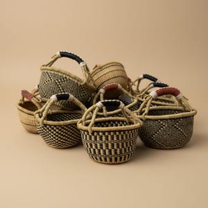 May include: A collection of small, round, hand-woven baskets made from natural fibers. Each basket features intricate black and tan geometric patterns and a single arched handle wrapped in either black or reddish-brown leather at the center.