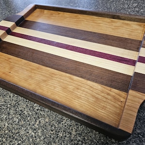 May include: A rectangular wooden serving tray with a dark brown border and handles. The tray's surface features alternating stripes of light, medium, and dark brown wood, with a thin purple stripe. The tray is designed for serving food or displaying items.