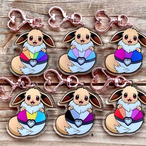 Może przedstawiać: Siedem kolorowych breloków z kreskówkowym Eevee trzymającym Poké Ball. Każdy Poké Ball ma inny schemat kolorystyczny reprezentujący flagę dumy LGBTQ+.
