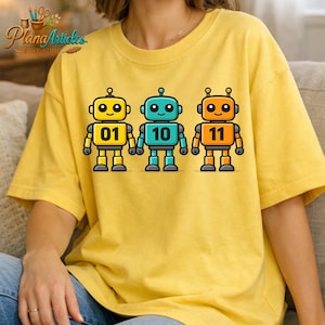 May include: Yellow t-shirt featuring three cartoon robots in yellow, teal, and orange. Each robot has a different number on its chest: 01, 10, and 11. The robots have antenna and are outlined in black.