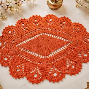 May include: An oval, orange crochet doily with a detailed openwork design. The doily features a central diamond pattern and a scalloped edge with decorative holes. This handmade piece adds a touch of vintage charm to any home decor.