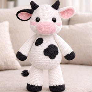 Op de afbeelding: Een handgemaakte gehaakte koeienknuffel. De witte koe heeft zwarte vlekken, hoeven en hoorns. De oren en de snuit zijn roze. De koe staat op een beige bank. Een schattig en knuffelbaar knuffeldier.