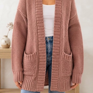 May include: A dusty rose open-front cardigan featuring a chunky knit texture, long sleeves, ribbed cuffs, a wide ribbed collar, and two large patch pockets on the front. The garment is worn over a white tank top and light blue denim jeans.