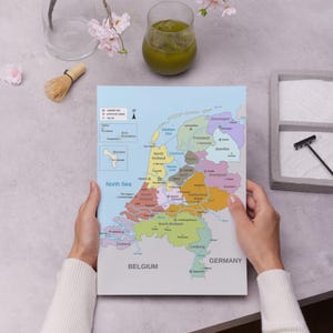 May include: A colorful map of the Netherlands, with each province distinguished by a different color. The map includes labels for cities, the North Sea, Belgium, and Germany. The map is held by a person.