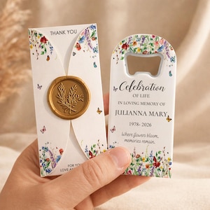 May include: A white card with a gold wax seal featuring a leaf design and a white bottle opener, both decorated with colourful wildflower and butterfly prints. The card reads THANK YOU and the bottle opener reads Celebration OF LIFE IN LOVING MEMORY OF JULIANNA MARY 1978-2026 Where flowers bloom, memories remain.