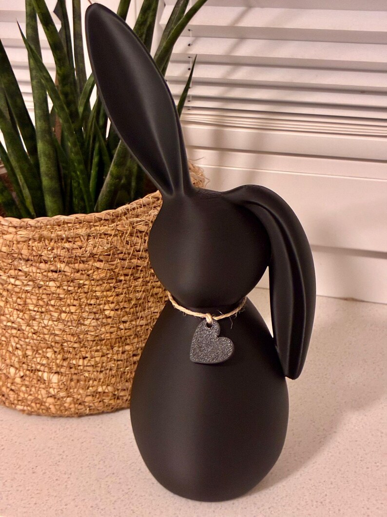 May include: A matte black bunny figurine with long ears and a heart-shaped pendant. The bunny is wearing a twine necklace with a small, textured heart. The figurine is a decorative item.