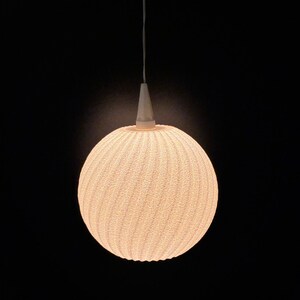Puede incluir: Una l&aacute;mpara colgante esf&eacute;rica con una superficie texturizada similar a un tejido. La luz es de un color blanco roto c&aacute;lido y cuelga de un accesorio c&oacute;nico blanco y un fino cable blanco sobre un fondo negro.