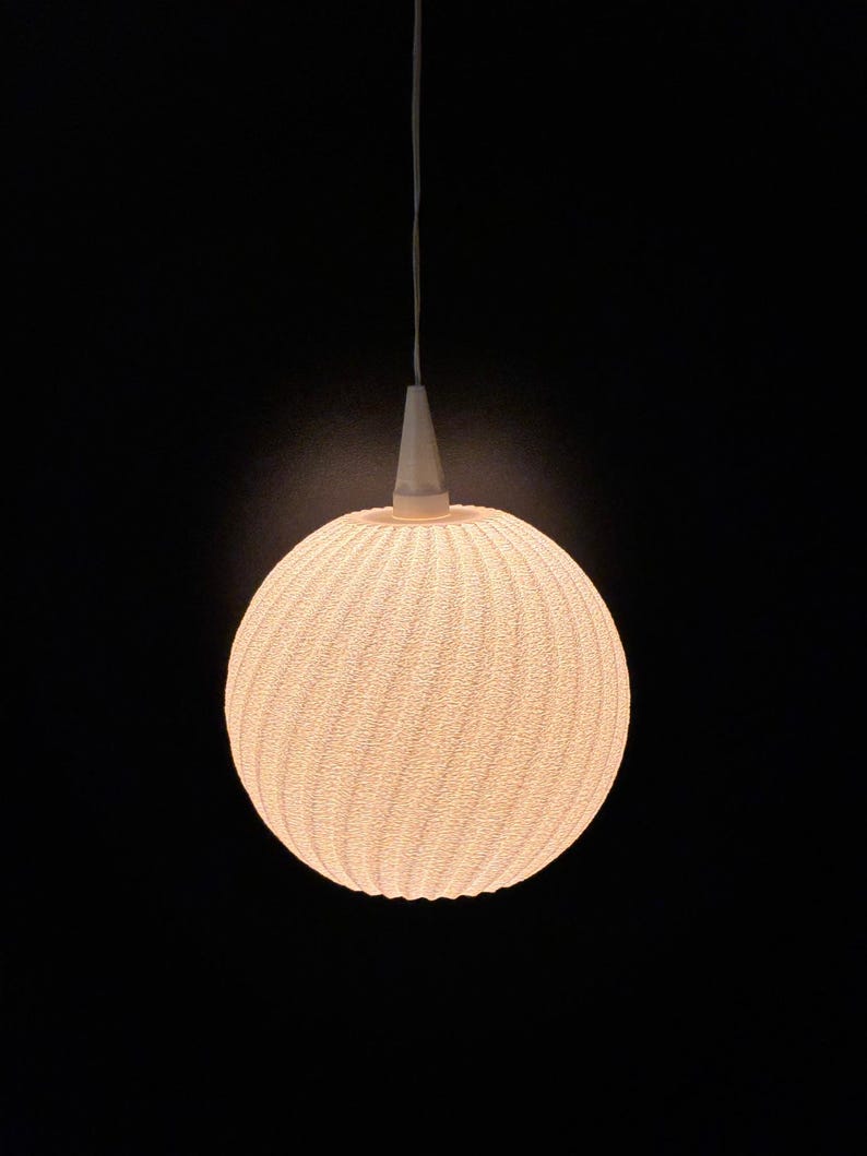 Puede incluir: Una l&aacute;mpara colgante esf&eacute;rica con una superficie texturizada similar a un tejido. La luz es de un color blanco roto c&aacute;lido y cuelga de un accesorio c&oacute;nico blanco y un fino cable blanco sobre un fondo negro.