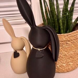 May include: Two decorative bunny figurines. One is beige, the other is black. Each has a heart-shaped tag hanging from a twine necklace. The bunnies have long ears and a smooth, egg-shaped body. A woven basket with a green plant is in the background.