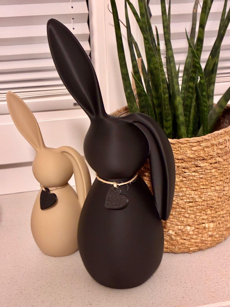 May include: Two decorative bunny figurines. One is beige, the other is black. Each has a heart-shaped tag hanging from a twine necklace. The bunnies have long ears and a smooth, egg-shaped body. A woven basket with a green plant is in the background.