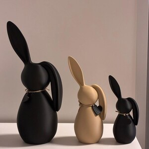 May include: Three decorative bunny figurines in black and tan. Each bunny has long ears and a rounded body. The bunnies are adorned with a small heart-shaped tag and twine around their necks. The bunnies are different sizes.