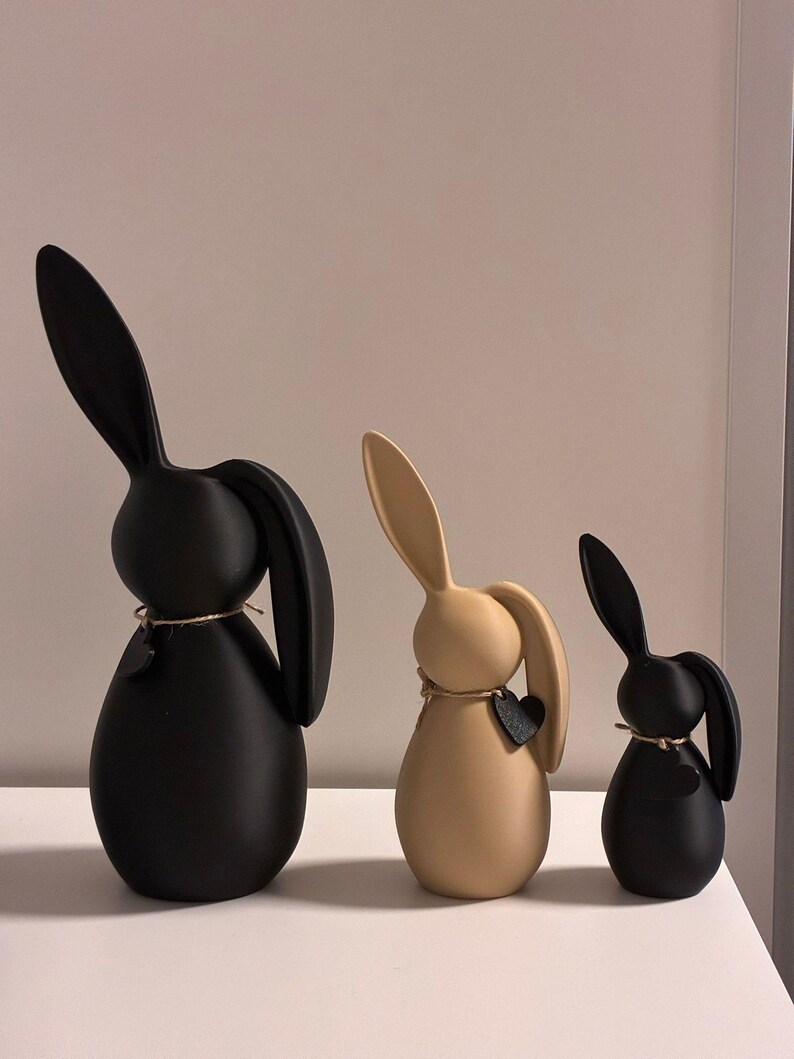 May include: Three decorative bunny figurines in black and tan. Each bunny has long ears and a rounded body. The bunnies are adorned with a small heart-shaped tag and twine around their necks. The bunnies are different sizes.