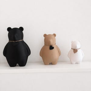 Pu&ograve; includere: Tre statuette a forma di orso in nero, beige e bianco, ognuna con un dettaglio a forma di cuore e una collana di spago. Le statuette sono esposte su una mensola bianca, mostrando un design minimalista.