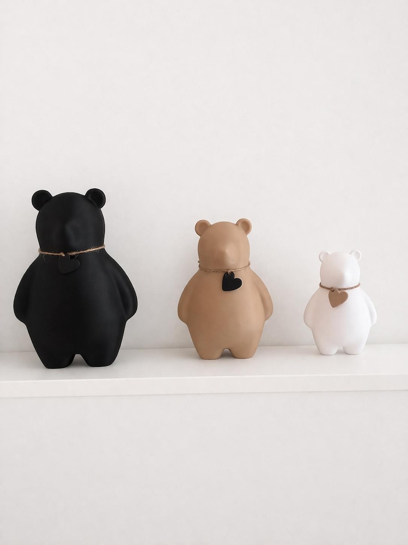 Pu&ograve; includere: Tre statuette a forma di orso in nero, beige e bianco, ognuna con un dettaglio a forma di cuore e una collana di spago. Le statuette sono esposte su una mensola bianca, mostrando un design minimalista.