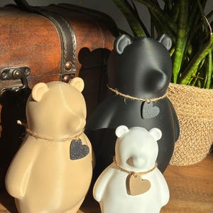 Pu&ograve; includere: Tre statuette di orso in beige, nero e bianco, ciascuna con un'etichetta a forma di cuore e una collana di spago. Gli orsi sono disposti su una superficie di legno, con un baule vintage e una pianta in vaso sullo sfondo.