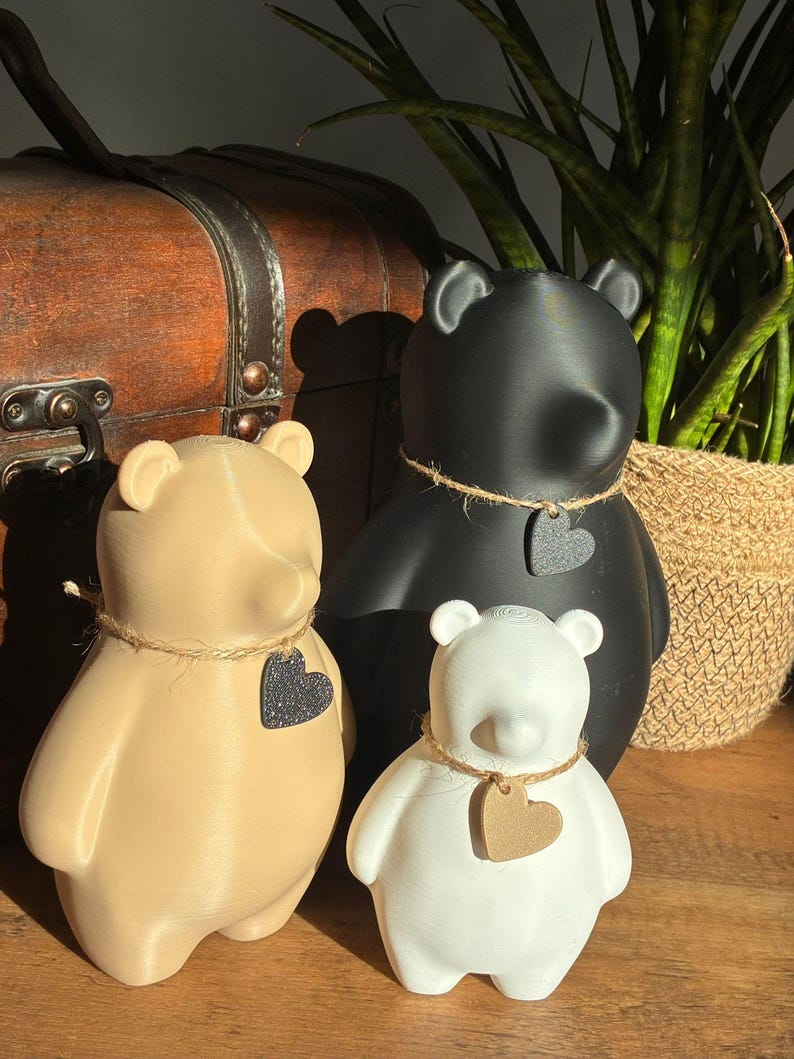 Pu&ograve; includere: Tre statuette di orso in beige, nero e bianco, ciascuna con un'etichetta a forma di cuore e una collana di spago. Gli orsi sono disposti su una superficie di legno, con un baule vintage e una pianta in vaso sullo sfondo.