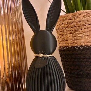 May include: A black decorative bunny figurine with long ears and a textured, ribbed body. The bunny has a small heart-shaped tag tied around its neck with twine. A gold-colored glass vase and a woven basket are in the background.