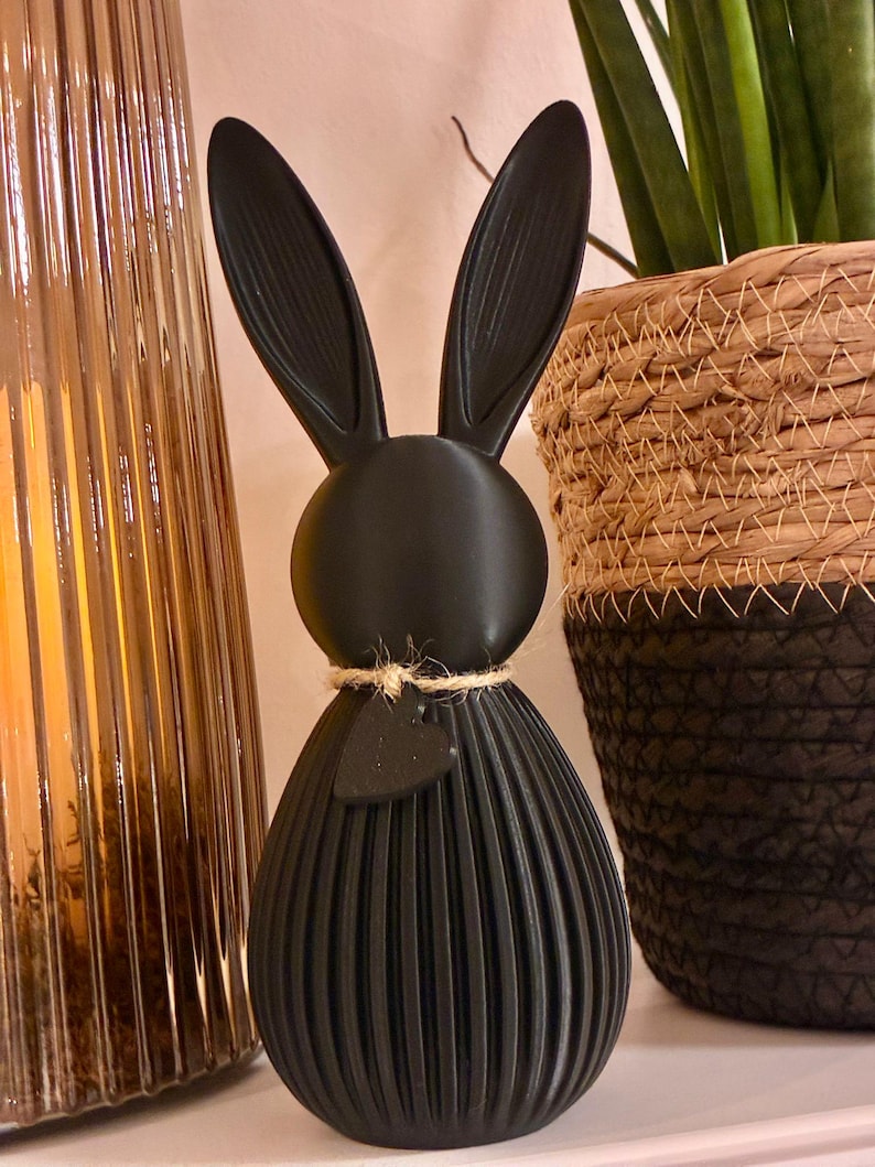 May include: A black decorative bunny figurine with long ears and a textured, ribbed body. The bunny has a small heart-shaped tag tied around its neck with twine. A gold-colored glass vase and a woven basket are in the background.
