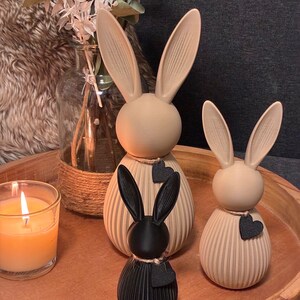 May include: Three decorative bunny figurines in a wooden tray. Two are beige with black heart accents, and one is black. A lit candle in a glass and a vase with dried flowers are also in the image. The bunnies have long ears.