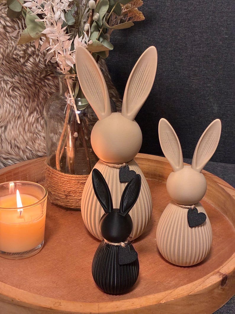 May include: Three decorative bunny figurines in a wooden tray. Two are beige with black heart accents, and one is black. A lit candle in a glass and a vase with dried flowers are also in the image. The bunnies have long ears.