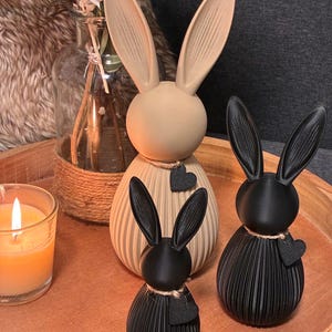 May include: Three decorative bunny figurines in beige and black, arranged on a wooden tray. The bunnies have long ears and are adorned with heart-shaped accents. A lit candle and a vase with dried flowers are also present.