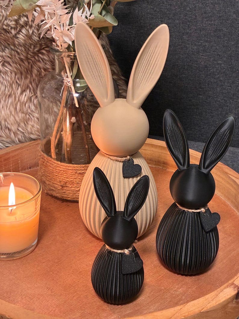 May include: Three decorative bunny figurines in beige and black, arranged on a wooden tray. The bunnies have long ears and are adorned with heart-shaped accents. A lit candle and a vase with dried flowers are also present.