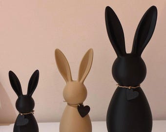 Può includere: Tre statuette decorative a forma di coniglietto pasquale di diverse dimensioni. I coniglietti sono neri e beige, con lunghe orecchie e un ciondolo a forma di cuore intorno al collo. I coniglietti sono su una superficie bianca.